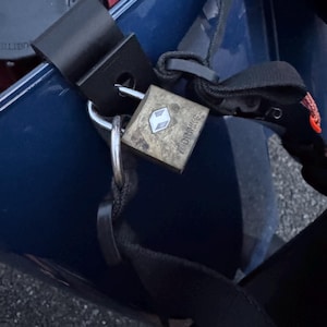 May include: A weathered, square padlock with the brand name "GRIMMS" is secured to a black strap. The lock is silver with a white diamond-shaped logo. The lock is attached to a black metal bracket. The background is a dark blue surface.
