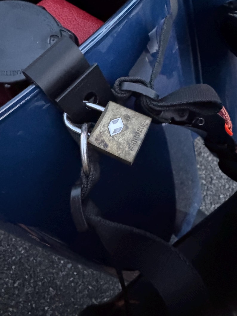 May include: A weathered, square padlock with the brand name "GRIMMS" is secured to a black strap. The lock is silver with a white diamond-shaped logo. The lock is attached to a black metal bracket. The background is a dark blue surface.