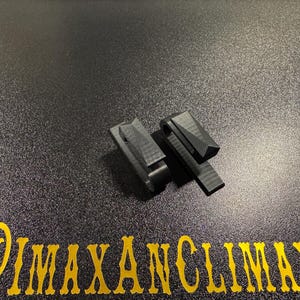May include: Two black, geometric-shaped clips are displayed on a textured, dark surface. The clips have a rectangular base with a hinged, angled top. The text "@IMAXANCLIMAX" is printed in gold at the bottom.