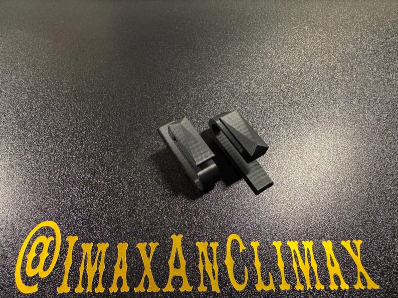 May include: Two black, geometric-shaped clips are displayed on a textured, dark surface. The clips have a rectangular base with a hinged, angled top. The text "@IMAXANCLIMAX" is printed in gold at the bottom.