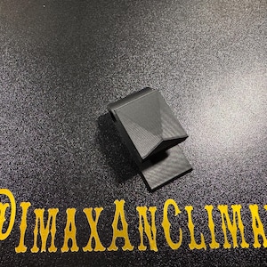 May include: A black, geometric-shaped object with a textured surface, possibly a clip or holder. The object is on a dark, speckled surface. The text "@IMAXANCLIMAX" is in gold lettering below the object.