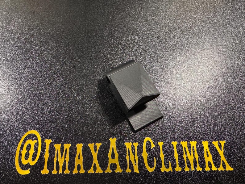 May include: A black, geometric-shaped object with a textured surface, possibly a clip or holder. The object is on a dark, speckled surface. The text "@IMAXANCLIMAX" is in gold lettering below the object.