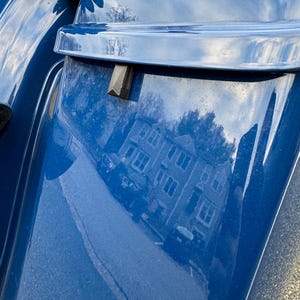 May include: Close-up of a section of a blue motorcycle. The glossy surface reflects a building and sky. A black rectangular piece is attached to the blue panel. The image is taken outdoors.
