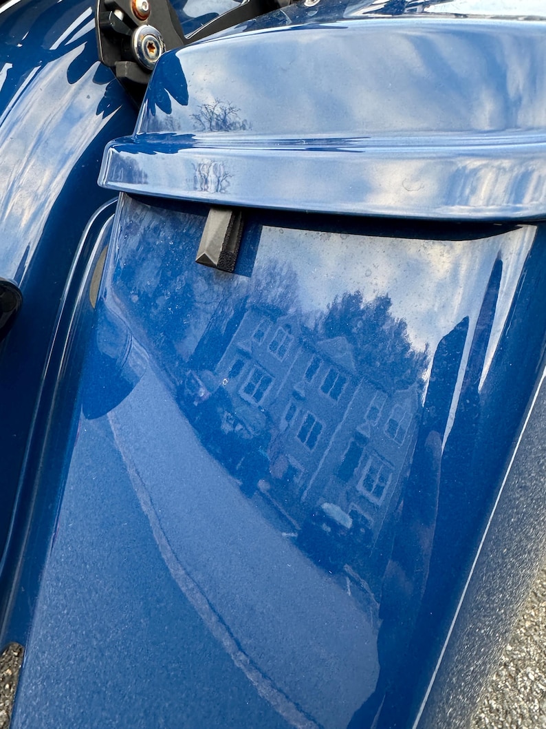 May include: Close-up of a section of a blue motorcycle. The glossy surface reflects a building and sky. A black rectangular piece is attached to the blue panel. The image is taken outdoors.
