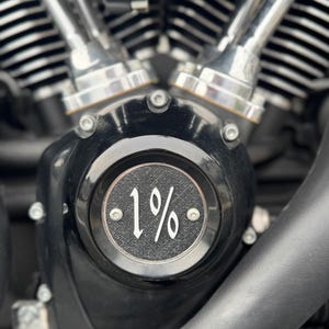May include: Close-up of a motorcycle engine with polished chrome cylinders and a black engine cover. The cover features the text "1%" in white script, surrounded by a textured border. The engine components are detailed and reflect light.