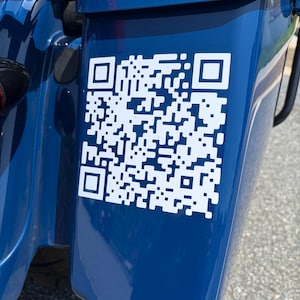 Puede incluir: Una pegatina de código QR blanca aplicada en el panel lateral azul brillante de una motocicleta. El gráfico cuadrado presenta un patrón de alto contraste de cuadrados blancos sobre la superficie azul oscuro del compartimento rígido.