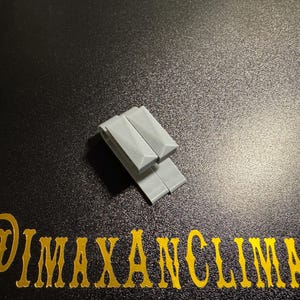 May include: Two light gray, rectangular objects are stacked on a dark, textured surface. The objects have a geometric design. Gold text at the bottom reads "@IMAXANCLIMAX".