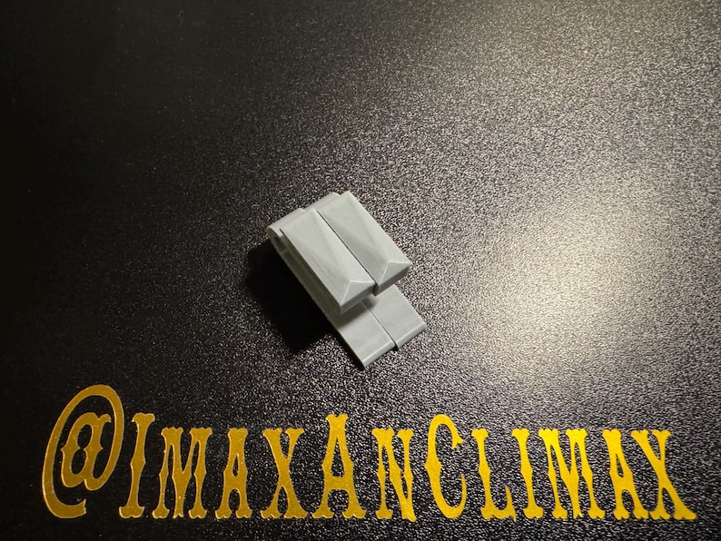 May include: Two light gray, rectangular objects are stacked on a dark, textured surface. The objects have a geometric design. Gold text at the bottom reads "@IMAXANCLIMAX".