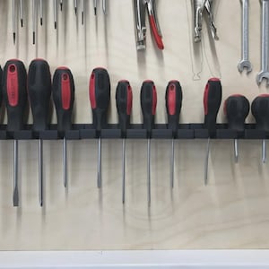 May include: A collection of screwdrivers with black handles and red accents, neatly organized on a black holder. The screwdrivers are of varying sizes and shapes, with silver metal shafts. Other tools, including wrenches and pliers, are visible in the background.