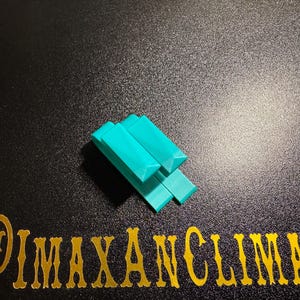 May include: A teal-colored, geometric puzzle piece composed of several rectangular blocks with angled edges. The blocks are arranged in a stacked configuration. The image also includes the text "@IMAXANCLIMAX" in gold lettering.