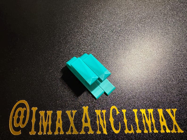May include: A teal-colored, geometric puzzle piece composed of several rectangular blocks with angled edges. The blocks are arranged in a stacked configuration. The image also includes the text "@IMAXANCLIMAX" in gold lettering.
