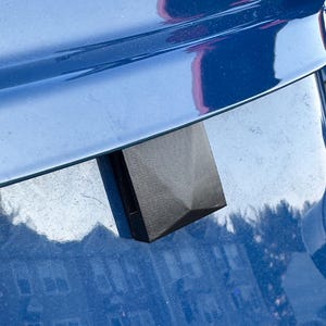 May include: A black, geometric-shaped object with a textured surface, possibly a car part, is attached to a shiny, blue surface. The object has a pyramidal top and a rectangular base. The blue surface reflects the surrounding environment.