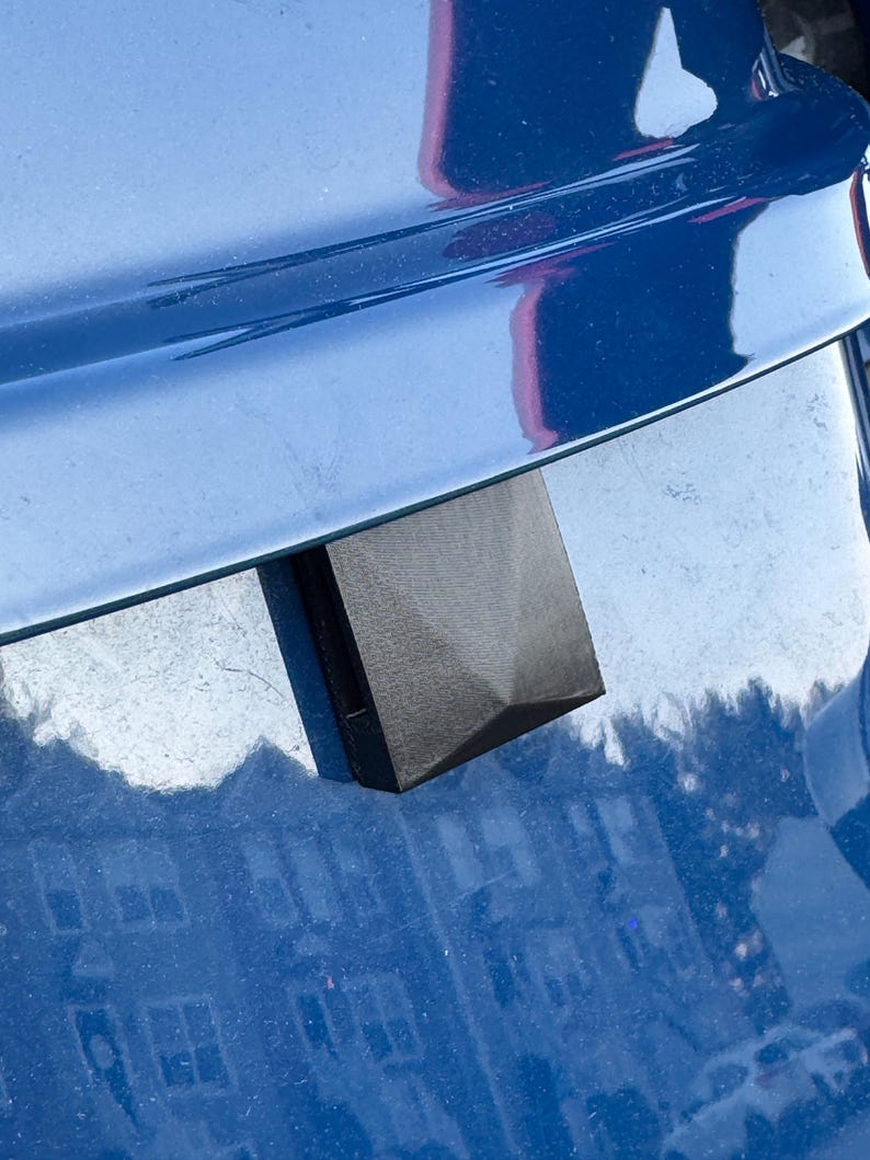 May include: A black, geometric-shaped object with a textured surface, possibly a car part, is attached to a shiny, blue surface. The object has a pyramidal top and a rectangular base. The blue surface reflects the surrounding environment.