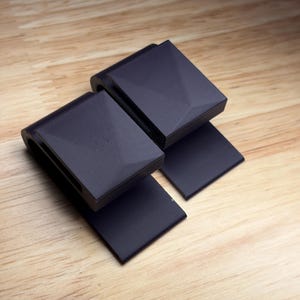 May include: Two matte black, U-shaped clips with a geometric, faceted top surface. Each clip features a flat, rectangular base extending from the curved body, constructed with a textured finish visible on the sides and smooth top panels.