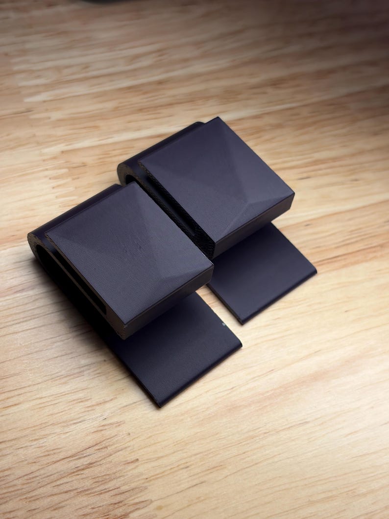 May include: Two matte black, U-shaped clips with a geometric, faceted top surface. Each clip features a flat, rectangular base extending from the curved body, constructed with a textured finish visible on the sides and smooth top panels.