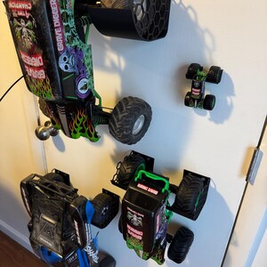 Puede incluir: Varios camiones monstruo de juguete, incluido un modelo "Grave Digger", est&aacute;n montados en una pared. Los camiones presentan dise&ntilde;os negros, verdes y azules con neum&aacute;ticos grandes. Tambi&eacute;n se ve un cami&oacute;n m&aacute;s peque&ntilde;o.