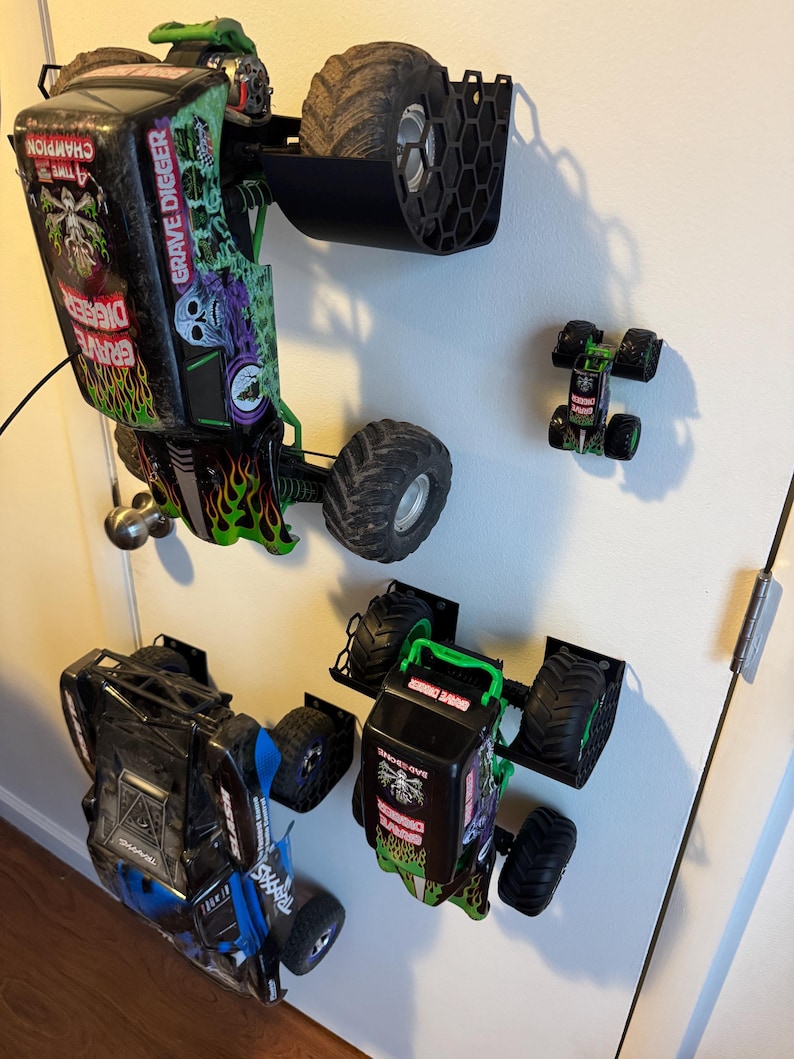 Puede incluir: Varios camiones monstruo de juguete, incluido un modelo "Grave Digger", est&aacute;n montados en una pared. Los camiones presentan dise&ntilde;os negros, verdes y azules con neum&aacute;ticos grandes. Tambi&eacute;n se ve un cami&oacute;n m&aacute;s peque&ntilde;o.
