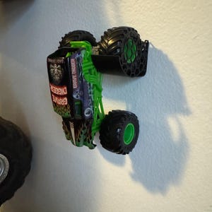 Puede incluir: Un juguete de cami&oacute;n monstruo Grave Digger verde y negro con neum&aacute;ticos negros grandes y llantas verdes. El cami&oacute;n tiene un chasis verde y las palabras "Grave Digger" son visibles en el lateral. El cami&oacute;n est&aacute; inclinado contra una pared blanca.