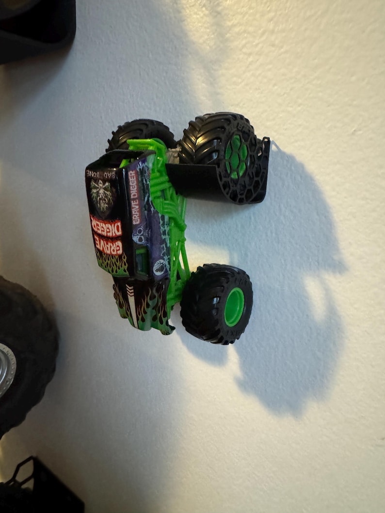 Puede incluir: Un juguete de cami&oacute;n monstruo Grave Digger verde y negro con neum&aacute;ticos negros grandes y llantas verdes. El cami&oacute;n tiene un chasis verde y las palabras "Grave Digger" son visibles en el lateral. El cami&oacute;n est&aacute; inclinado contra una pared blanca.