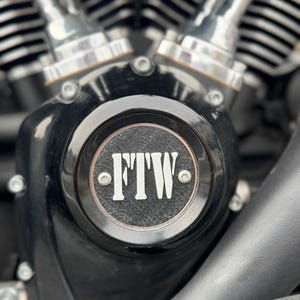 May include: Close-up of a motorcycle engine with a black cover. The cover has a circular emblem with the letters "FTW" in white. The engine components are silver and chrome, with visible bolts and a textured surface.