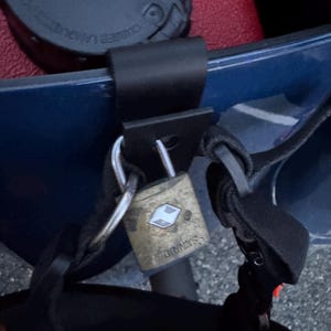 May include: A brass-colored padlock with the brand name "Brinks" is secured to a black strap. The lock is attached to a blue surface, with a black strap and a red surface visible in the background.