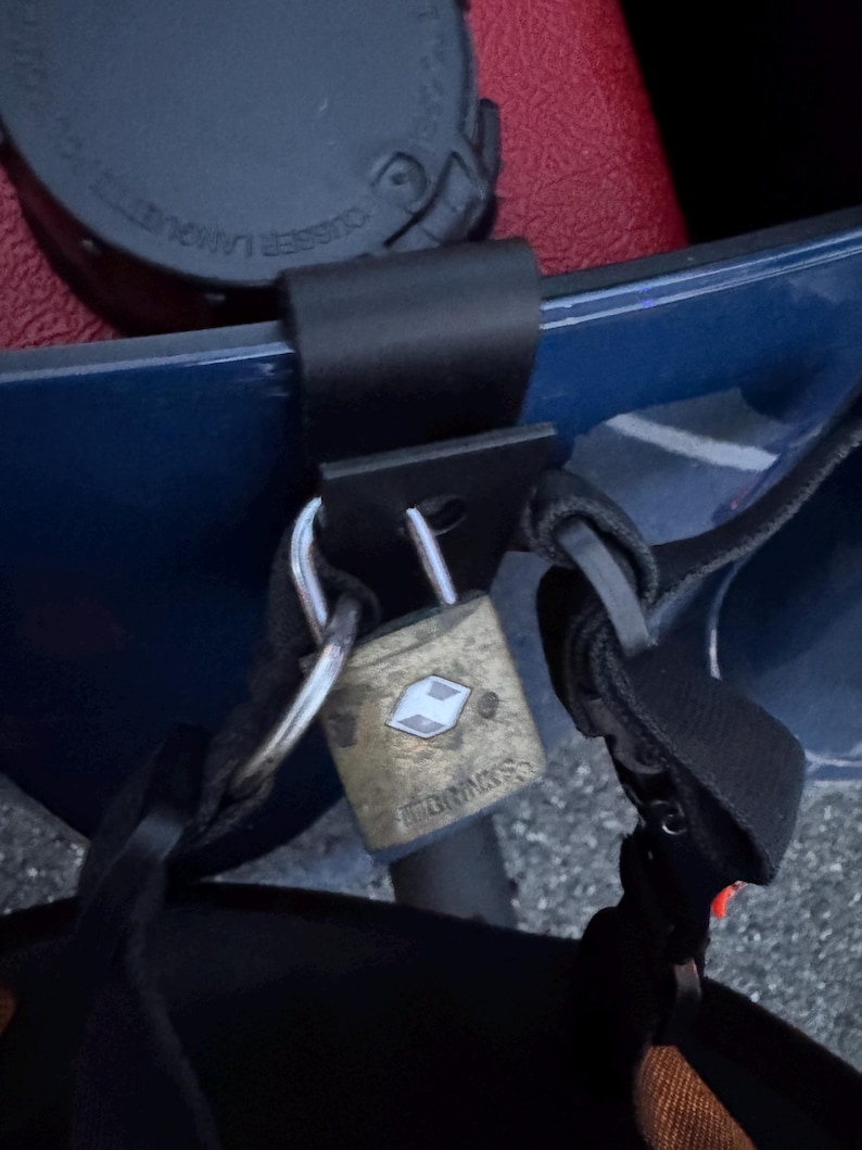 May include: A brass-colored padlock with the brand name "Brinks" is secured to a black strap. The lock is attached to a blue surface, with a black strap and a red surface visible in the background.