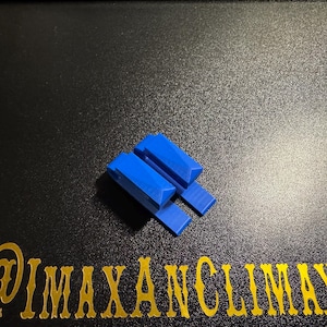 May include: A small, bright blue, geometric object with a rectangular base and two angled, rectangular pieces on top. The object is on a black surface with gold text that reads "@IMAXANCLIMAX".