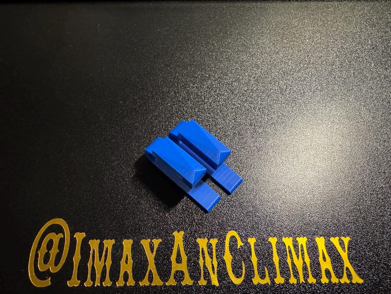 May include: A small, bright blue, geometric object with a rectangular base and two angled, rectangular pieces on top. The object is on a black surface with gold text that reads "@IMAXANCLIMAX".