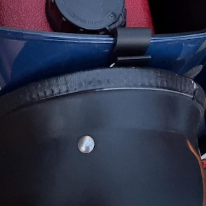 May include: Close-up of a black object with a silver-colored, round, metal stud. A black strap is attached to a blue surface. A black cap is visible in the background, resting on a red surface.