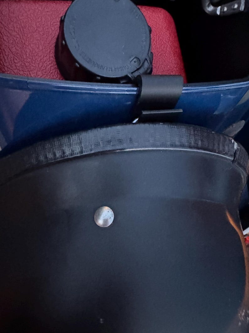 May include: Close-up of a black object with a silver-colored, round, metal stud. A black strap is attached to a blue surface. A black cap is visible in the background, resting on a red surface.