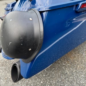 May include: Close-up of a motorcycle's rear section, featuring a glossy blue body and a black exhaust pipe. A black, rounded component with rivets is attached to the side, and a red reflector is visible.