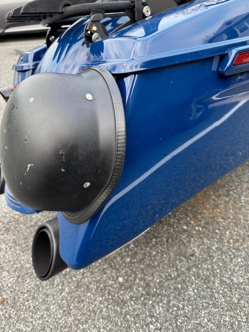 May include: Close-up of a motorcycle's rear section, featuring a glossy blue body and a black exhaust pipe. A black, rounded component with rivets is attached to the side, and a red reflector is visible.