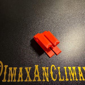 May include: Bright red plastic clips arranged in a stack on a black surface. The clips have a rectangular shape with a small triangular cut-out at the top. The text "@IMAXANCLIMAX" is in gold lettering.