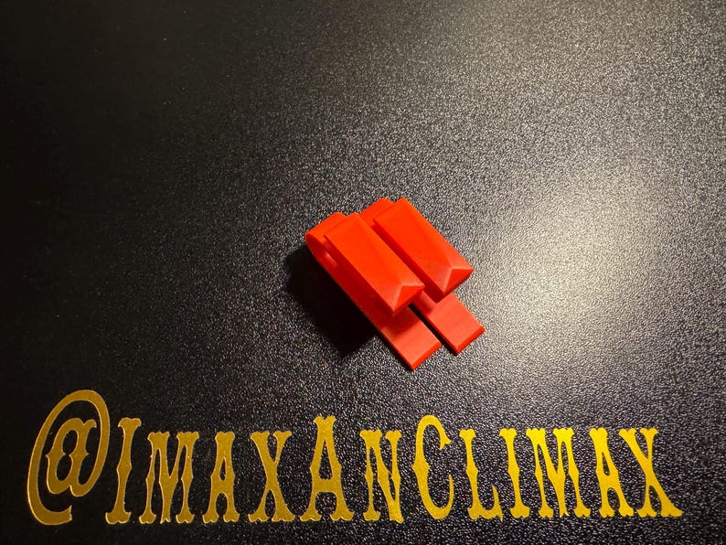 May include: Bright red plastic clips arranged in a stack on a black surface. The clips have a rectangular shape with a small triangular cut-out at the top. The text "@IMAXANCLIMAX" is in gold lettering.