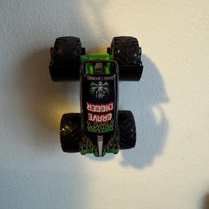 Puede incluir: Un juguete de cami&oacute;n monstruo Grave Digger negro y verde con neum&aacute;ticos negros grandes. El cami&oacute;n presenta una jaula antivuelco verde y las palabras "Grave Digger" en llamas verdes y rojas. La parte superior del cami&oacute;n dice "Bad Bone".