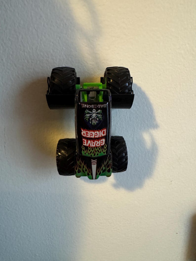 Puede incluir: Un juguete de cami&oacute;n monstruo Grave Digger negro y verde con neum&aacute;ticos negros grandes. El cami&oacute;n presenta una jaula antivuelco verde y las palabras "Grave Digger" en llamas verdes y rojas. La parte superior del cami&oacute;n dice "Bad Bone".