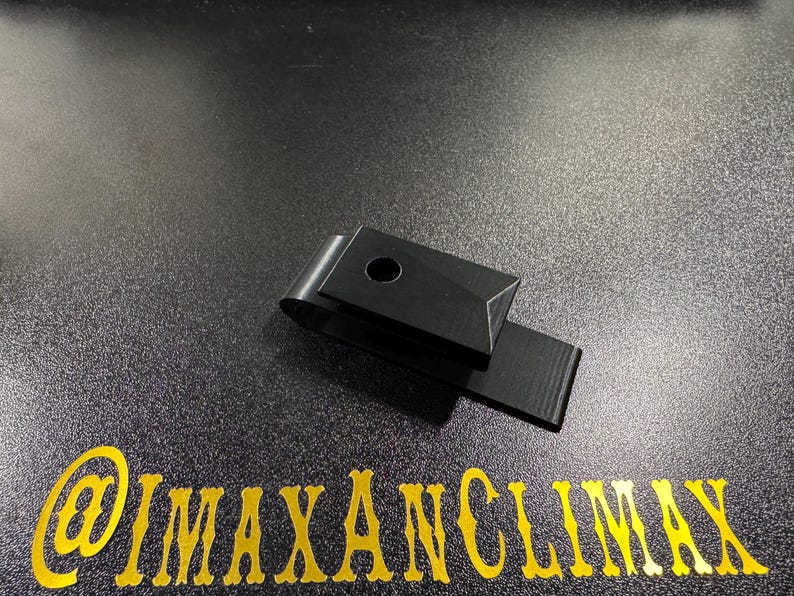 May include: A black metal money clip with a rectangular design and a circular hole. The clip is shown on a dark surface with the gold text "@IMAXANCLIMAX".