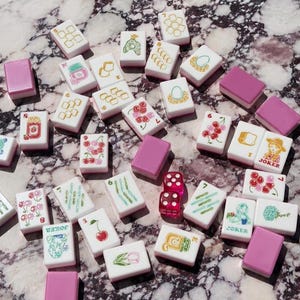 May include: A collection of white mahjong tiles featuring colorful illustrations of fruits, flowers, and characters, alongside solid pink tiles and two translucent pink dice. The tiles display various symbols, including cherries, tulips, and the word Joker.