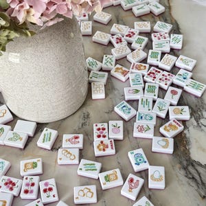 May include: A collection of white rectangular Mahjong tiles scattered across a surface, featuring colorful illustrations of flowers, animals, food, and the word Joker. A speckled ceramic vase with pink hydrangea flowers sits nearby.