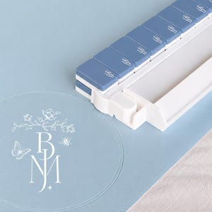 May include: A light blue pill organizer with seven compartments, each labeled with a small icon. The organizer sits on a matching blue surface, next to a round mat with a floral and butterfly design and the initials "BNJ".