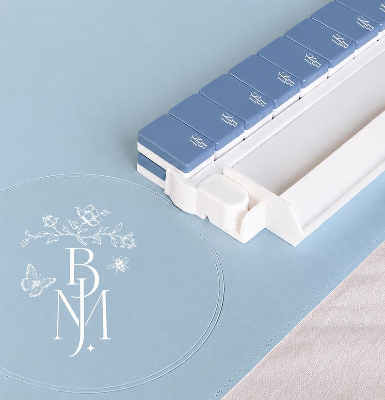 May include: A light blue pill organizer with seven compartments, each labeled with a small icon. The organizer sits on a matching blue surface, next to a round mat with a floral and butterfly design and the initials "BNJ".