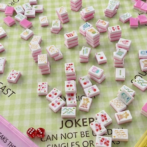 Pu&ograve; includere: Una collezione di tessere Mahjong rosa e bianche con varie illustrazioni, tra cui ciliegie e un jolly, sparse su una superficie a scacchi verdi. Il set include dadi e un astuccio rosa. Il testo sull'astuccio recita "BREAKS THE WALL AND ST".