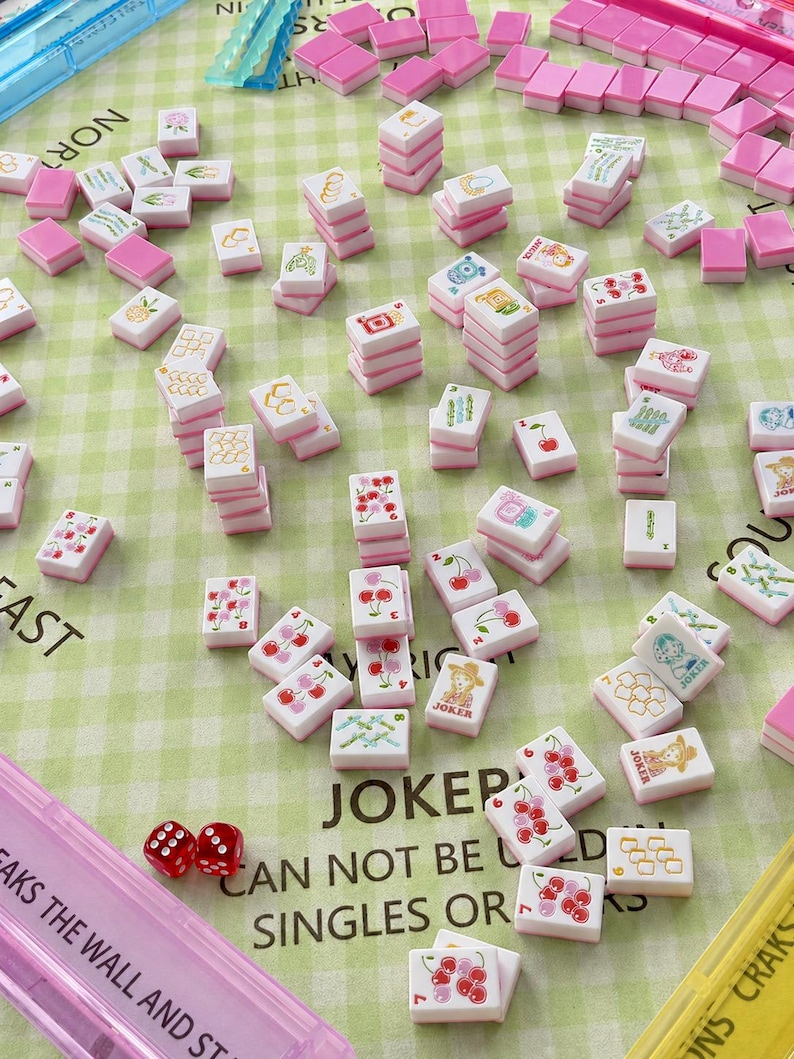 Pu&ograve; includere: Una collezione di tessere Mahjong rosa e bianche con varie illustrazioni, tra cui ciliegie e un jolly, sparse su una superficie a scacchi verdi. Il set include dadi e un astuccio rosa. Il testo sull'astuccio recita "BREAKS THE WALL AND ST".