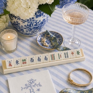 May include: A still life arrangement featuring a Mahjong tile set, a champagne glass, and a floral napkin with embroidered initials. A gold bracelet and sunglasses are also present. The scene is set on a blue and white striped tablecloth, with a vase of white and blue hydrangeas.