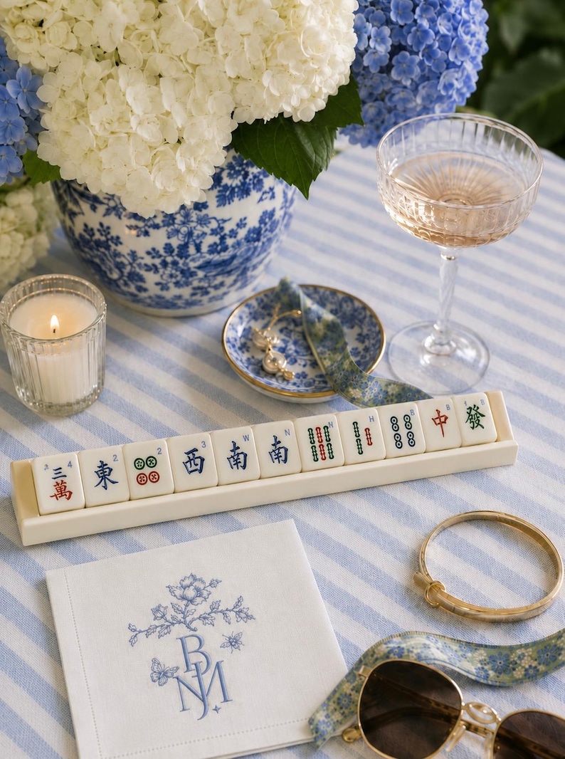May include: A still life arrangement featuring a Mahjong tile set, a champagne glass, and a floral napkin with embroidered initials. A gold bracelet and sunglasses are also present. The scene is set on a blue and white striped tablecloth, with a vase of white and blue hydrangeas.