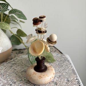 May include: A decorative sculpture featuring a round, speckled beige stone base topped with a dark brown cylindrical mount. Thin gold wires extend upward, each adorned with various buttons in shades of brown, cream, and white, along with clear beads.