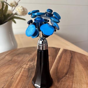 May include: A decorative floral arrangement featuring a black glass vase with a silver-colored top, holding a bouquet of blue buttons. The buttons vary in shades of blue and are attached to wire stems.