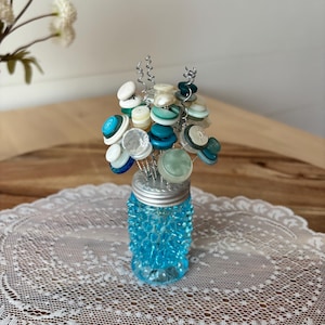 May include: A decorative arrangement featuring a cluster of assorted white, teal, and blue buttons attached to silver wire stems, displayed inside a small, textured, bright blue glass jar with a silver-colored metal lid.