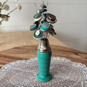 May include: A decorative object featuring a teal ceramic base with a silver-toned metal top. Thin, coiled silver wires extend upward from the base, each topped with a variety of round buttons in shades of teal, white, gray, brown, and black.