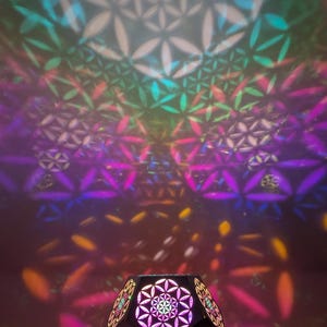 May include: A geometric, multi-faceted lamp with a floral pattern, projecting colorful light patterns onto the wall. The lamp's design includes a base and is illuminated with pink, yellow, and blue hues. The image includes the text "Amberlights Handmade Sensory Art".